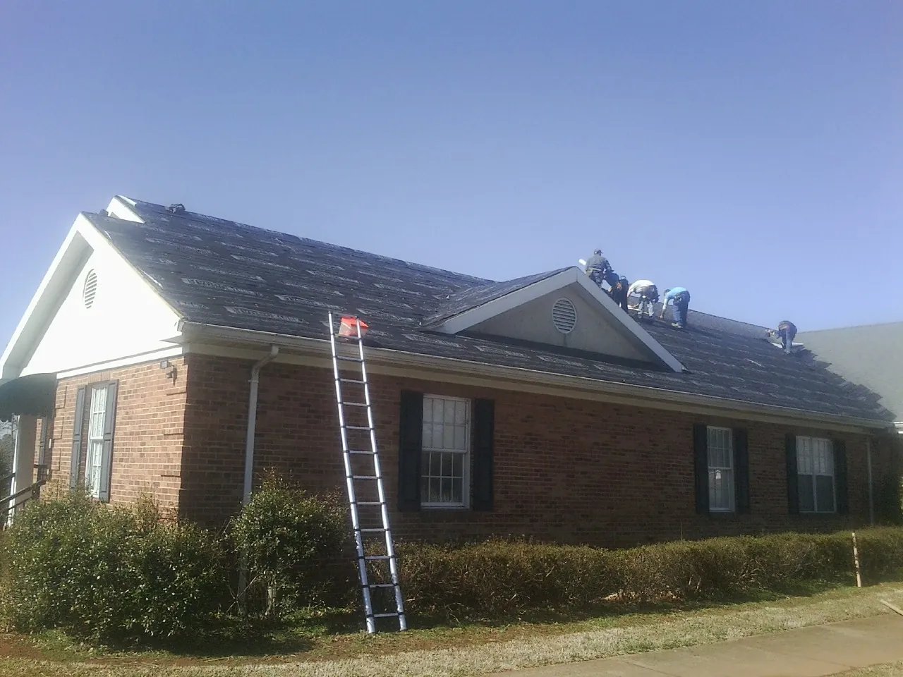 Crew tearing shingles off a brick ranch roof with a ladder set — Metal Roof Repair work by our crew in Guernsey