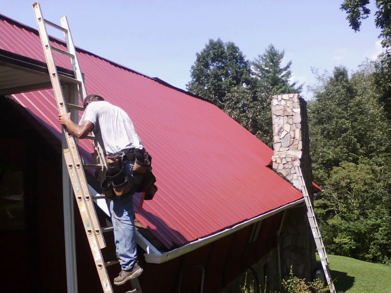 Roofer on a ladder beside a red metal roof and stone chimney — Soffit & Fascia Repair by Roofing Brothers in Guernsey